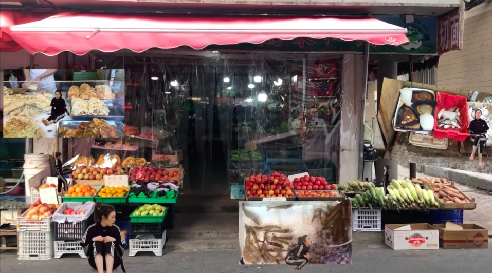 A still from a video showing a grocery store and food piled up outside it on display, for sale. A figure sits in front of one of the piles looking right.